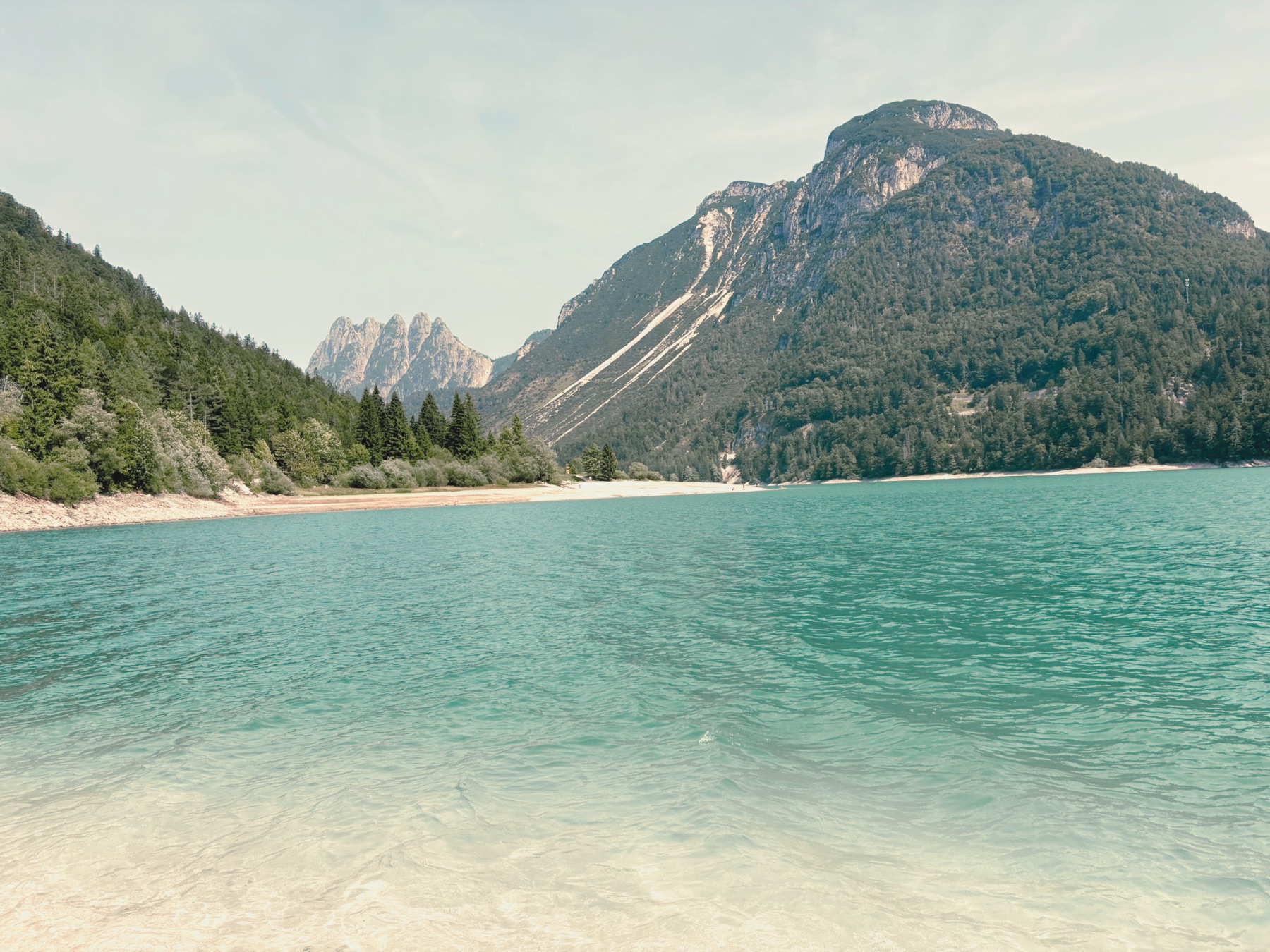 Ein klarer Bergsee in Türkis, dahinter bewaldete Hänge und Felsen